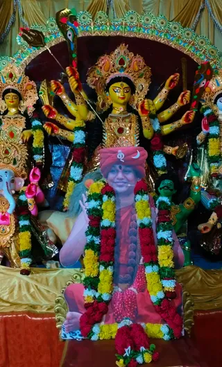 A large cutout photograph of a smiling figure in a red-orange turban with a crescent moon emblem and red-orange robes, garlanded with marigold and red flower garlands, placed seated on a gold throne in front of a deity murti. The deity is a multi-armed yellow goddess with a gold crown and red jewelled ornaments, holding weapons including a trident, surrounded by an elephant-headed Ganesha murti, a green figure and other murtis in a decorated arch with gold drapery. The cutout is a printed picture, not a physical person. Decorated festival puja display.