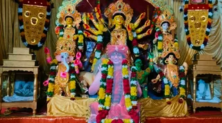 A decorated shrine with a multi-armed goddess murti in gold and red, flanked by Ganesha in gold and figures in yellow and green. Marigold and rose garlands drape the murtis. In front, a framed-looking figure in a pink and orange robe with a pink turban and crescent emblem sits draped in garlands, appearing to be a sculpted or printed portrait rather than a physical person. Small framed photographs appear on wooden side stands, and golden banners with garlands hang at the top corners.