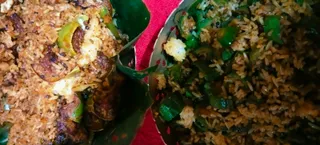 Close overhead photo of two portions of spiced rice served on green leaf plates or cups over a red cloth. The rice is mixed with sliced green peppers, pieces of soft white paneer or cheese, and darker fried pieces, with mustard seeds and curry leaves visible. No people are present.