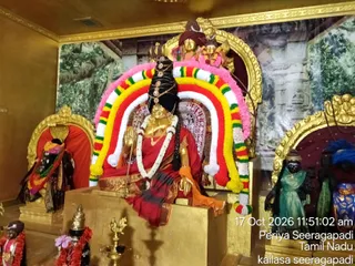 Interior temple shrine showing a golden deity murti of a goddess seated on a raised golden peetha, draped in a red sari with gold border and ornate gold jewellery, with a rudraksha-style headdress and a large crescent arch of woven garlands in red, yellow, green, white and pink framing her. Smaller shrines flank her: at left a dark figure in a red-lined niche wearing a black and garland-draped shawl, and at right a dark figure in green cloth beside a blue and gold-patterned pedestal. Golden lamps and a decorated kalasha-style brass lamp sit in the foreground near small garlanded figures, and a painted backdrop shows a forest scene with stone architecture.