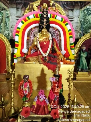 A golden deity murti seated on a gold pedestal under a colourful floral arch of red, yellow, white and green garlands. The figure wears a red and orange sari, a white jasmine garland, gold necklaces, and has a tall black hair crown adorned with gems; it holds a trident raised in one hand. Smaller dark deity figures in red garments and marigold garlands stand at the base, with hibiscus flowers placed before them. Brass oil lamps flank the pedestal. A standing green-clad figure appears in the background niche. The timestamp and place caption 'Periya Seeragapadi, Tamil Nadu, kailasa seeragapadi' are overlaid at lower right. No living people are visible.