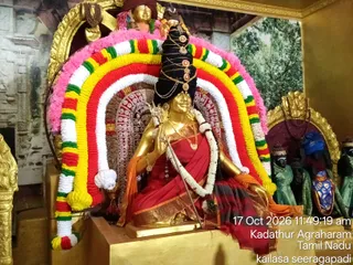 A golden deity murti of a goddess, seated in a shrine decorated with a multicolour garland arch of red, yellow, green and white flowers with pink edging. The murti wears a red and black sari with gold border, a crown-like black headdress with gold ornaments, a white flower garland, gold necklaces and bangles, and holds a trident. Two dark-skinned attendant figures in green robes stand at the right, and a painted tree backdrop with gold-framed panels surrounds the altar. A timestamp and location caption appear at lower right.