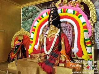 A photograph inside a temple shrine showing a golden deity murti of Goddess Durga seated in a red sari with gold border and an orange shawl, holding a trident-like staff in one hand and a gold hand raised in blessing. The murti wears a tall black crown with gold ornaments and pearl strands, a white jasmine garland, and a gold necklace. Behind her is a large arch of marigold-like garlands in yellow, red, green, white and pink. To the left, a smaller dark murti in a red cloth with a garland sits on a gold throne. The walls are gold with a painted forest backdrop. A timestamp reads 17 Oct 2026 11:49:11 am with the location Kadathur Agraharam, Tamil Nadu. No physical people are visible.