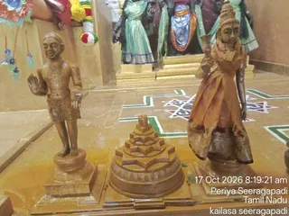 A photograph inside a temple shrine showing three golden-brown painted statues on a yellow floor with green and red geometric kolam lines. On the left stands a bare-chested male figure with one hand raised in blessing, wearing short dhoti-style garments. In the centre sits a stepped sri yantra-like meru or shrine-form object. On the right stands a female figure in an orange sari holding a ceremonial lamp-like object. Behind them, dark figures in green and blue saris stand on a golden platform, and garlands with red, green and white flowers hang at the upper left. A timestamp overlay reads 17 Oct 2026 8:19:21 am.