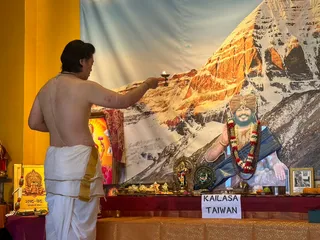 A shirtless man in white dhoti with gold border stands at left, raising a small oil lamp toward an altar in a puja. Behind him is a large backdrop of a snow-covered mountain face lit orange by sunlight. On the altar, cut-out or printed portrait of a bearded man wearing an orange-gold turban, rudraksha-style red-and-white flower garland, and blue shawl, with hands raised in blessing; a framed photo sits at right. Trays of fruit, flowers, brass vessels and small deity figures are arranged on a draped table with a white sign reading KAILASA TAIWAN. A yellow wall and a Shabd-Ved placard appear at left.