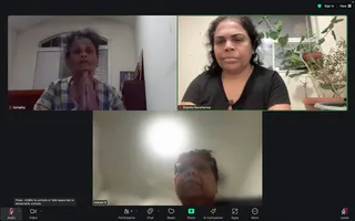 A screenshot of a Zoom video call with three video tiles. Top left, a woman in a blue and white plaid shirt sits with her palms pressed together in anjali mudra in front of a white wall, arched window and a small shrine with a lamp. Top right, a woman with dark curly hair in a black top and a thin gold necklace sits beside potted plants and a window. Bottom centre, a woman in glasses is seen from below in a brightly lit room. The Zoom control bar along the bottom shows Audio, Video, Participants 3, Chat, React, Share, AI Companion, Apps, More and Leave. Tile labels read komathy, Shanthi Ravishankar and Indrani S.