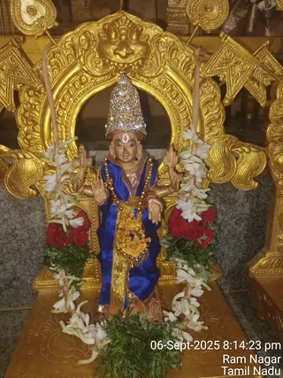 A photograph of a four-armed deity murti standing before a gilded arch in a temple shrine. The figure wears a tall crown encrusted with sparkling stones, a red tilak on the forehead, a royal-blue silk garment with gold border, a gold waist sash and a gold beaded chain. Two white jasmine garlands with red rose garlands hang on either side, and green leaves are placed at the base. Two tall candles stand beside the figure. The timestamp reads 06-Sept-2025 8:14:23 pm, Ram Nagar, Tamil Nadu. No physical people are visible.