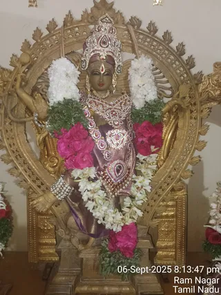 A photograph of a decorated deity murti in a golden prabhavali-style arch, showing a figure in a purple and gold silk garment with rhinestone and red-stone jewelled ornaments, a jewelled crown, and pearl bangles. Garlands of white jasmine-like flowers, pink roses and green leaves hang across the figure. The deity is set on a golden pedestal against a cream wall, with partial garlands visible at the edges. No people are visible. Timestamp overlay reads 06-Sept-2025 8:13:47 pm, Ram Nagar, Tamil Nadu.