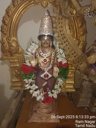 A photograph of a standing metal murti of a deity, decorated for worship and placed on a wooden table before an ornate gilded arch. The figure wears a tall crown encrusted with silver-white stones, a purple garment with gold border, a large red and green gemstone necklace and matching chest piece, and bangles. White jasmine garlands and red roses hang around the figure, and its right hand is raised in abhaya mudra. Behind it, a gilded deity figure and an ornate pedestal are partly visible. Timestamp text reads 06-Sept-2025 8:13:33 pm, Ram Nagar, Tamil Nadu.