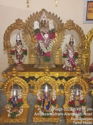 A photograph of a temple shrine displaying metal murti images of Shiva as Nataraja and associated deities, dressed with white jasmine garlands and red flower garlands. The central golden arch holds a standing figure with a silver crown and ornate jewellery, flanked by two figures with silver crowns; the figures on the right wear pink-purple silk and the one on the left wears a purple garment. Three smaller figures in golden arched niches on the lower tier wear silver crowns, blue garments and garlands. Gold swastika ornaments hang on the wall above, and a framed picture is partly visible at the top right. A timestamp reads 06-Sept-2025 8:13:21 pm, Ambasamudram-Alangulam Road, Kuthapanjan, Tamil Nadu. No people are visible.
