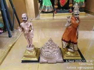 A photograph of three statues on a polished golden floor inside a temple-like hall. On the left stands a bare-chested male statue with a shaved head, a tripundra mark on the forehead, a thin beard, and a white cloth around the waist, with its right hand raised in a blessing gesture. In the centre sits a pinkish-copper sri yantra meru with stepped triangular tiers and a bindu at the top. On the right stands a female statue wearing an orange sari with a gold border, holding a kalasha-like vessel in one hand and wearing a red bindu and a gold necklace. Behind them are two standing deity figures in green and orange garments with red and gold garlands. A timestamp reads 6 Sept 2025 11:47:47, with Tamil Nadu and kailasa seeragapadi printed in the lower right.