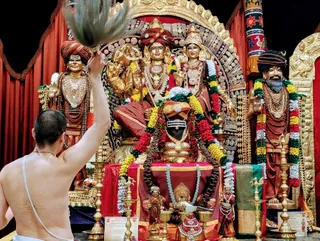 A photograph of a shaved-headed, bare-chested priest in a white dhoti and sacred thread, raising a white fan or chamara over a decorated altar. The central deity murti is a seated golden figure with a red turban, ornate jewellery and a crown-like headdress, flanked by a second golden figure in a crown and jewelled necklaces. Two standing sage statues in red and saffron robes, with rudraksha malas and gold necklaces, stand on either side. The black Shiva linga at the base is dressed in a red cloth and garlands of red, yellow and white flowers, with brass oil lamps and a small offering vessel in front. Red curtains and a decorated arch frame the scene.