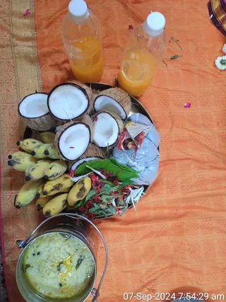 Top-down photo of a ritual offering on an orange and gold bordered silk cloth. A round metal tray holds halved white coconuts, a bunch of small yellow bananas, a leaf-wrapped packet, red and white flowers and green leaves. Two plastic bottles of orange liquid stand at the top, and a steel tiffin carrier holds a pale yellow rice dish with curry leaves. Scattered petals and a flower lie on the cloth. No people are visible.
