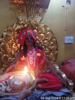 A seated figure in red garments and a red head wrap decorated with silver ornaments sits before an ornate gold cobra-hood backdrop. The figure wears a red shawl, a white beaded necklace and a garland of marigolds and pink flowers, and holds an offering tray of flowers and white material with a lit oil lamp flame. A wall switchboard and a red cushion are visible at right. The photo carries a timestamp of 06-Sep-2024 9:11:09 pm.