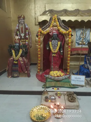 Indoor shrine photograph with gold-painted walls and a gold canopy. At left, a standing statue of a smiling man in a red-and-orange turban, saffron-toned draped garment, and a long garland of white and multicoloured flowers with beaded necklaces stands beside a dark stone elephant-headed deity seated on a pedestal, draped in red and striped silk and garlanded with yellow marigolds. At centre, a dark stone deity murti stands within a golden arched niche framed by orange and yellow marigold garlands, wearing a red cloth, ornate jewelled collar and a pink turban-like headdress. In front sits a brass plate heaped with yellow flowers, and a brass bell, brass trident, brass bowls and small oil lamps are arranged on a cloth. A framed black-and-white photo with a trident hangs at right, and a sign reads KAILASA THIRUCHENGODU ERODE. A timestamp reads 07 Sep 2024.