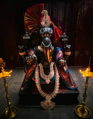A photograph of a dark-stone murti of Ganesha seated on a black pedestal against a dark red drape. The deity wears a red and gold headdress with a jewelled ornament, gold necklaces and a large layered gold-tone garland with a pendant, and a blue and red-gold silk cloth draped over his arms. Two brass oil lamps with lit wicks stand on either side. No physical people are visible.