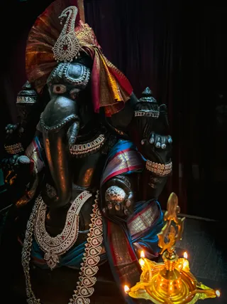 A dark-hued Ganesha murti is the main subject, seated and draped in a magenta and gold silk cloth with a blue and red border and wearing a jewelled crown-like headdress with red stones. Its ears, trunk and multiple arms are adorned with gold bracelets and layered jewelled necklaces set with red and green stones. Beside it at lower right stands a golden oil lamp with several lit flames on a lotus-shaped base, against a dark curtained background. No people are visible.