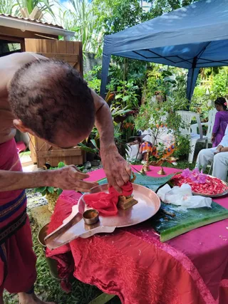 Outdoor puja in a garden under a blue pop-up canopy. A bare-chested priest in red dhoti with a dark striped waist cloth leans over a copper tray, wiping a small brass figure with red cloth. A pink table cover, banana leaves, a tray of red petals with purple flowers, brass oil lamps and a small bowl of kumkum sit on the table. Seated people in white and a child in purple watch from the background among tropical plants. No identification of a person is made from appearance; the priest is shown performing ritual cleansing.