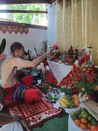 A photograph of a puja at a home shrine. A shirtless man with short grey hair sits cross-legged on a red and cream patterned mat, wearing a red dhoti with a striped border and holding a small ritual item toward a lit brass lamp. In front of him stands a copper kalasha pot, brass oil lamps, small deity murtis, white and pink flowers, red ginger blossoms, a steel plate of green leaves, a bottle, and a bowl of grapes, bananas, oranges and mangoes on the floor. Yellow curtains and a white cloth cover the altar table. Part of another person's arm and hand is visible at the lower left edge.