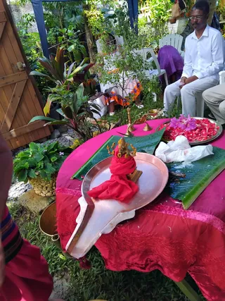 Outdoor ritual setup in a garden with lush tropical plants. In the foreground, a brass Ganesha murti wrapped in a red cloth sits on a large copper abhishekam plate, with a pink-draped table holding a banana leaf, a tray of red flower petals with purple blossoms, brass bells and an oil lamp. A seated man in a white shirt and white trousers watches the ritual from a white plastic chair, and a partially visible person in purple sits nearby. A wooden gate stands at left. The scene suggests a puja with abhishekam offerings.