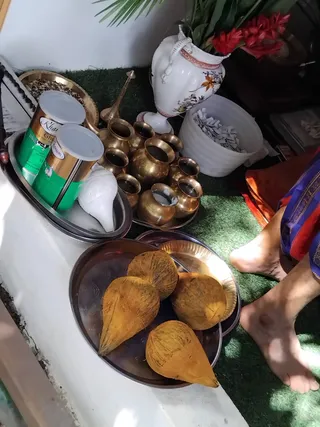 A photograph of puja preparations on green artificial grass beside a white ledge. A steel tray holds several peeled brown coconuts, and a second tray holds two tins of ghee labelled with green and gold packaging, a white conch shell, and a bowl of grains. A cluster of brass kalasha-style vessels and a brass lamp stand sit nearby, with a white ceramic vase of red flowers and a white bucket of folded paper. Bare feet and an orange and blue cloth are visible at the right edge, but no person's face is shown.