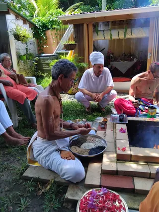 Outdoor homa at a garden home temple. A shirtless older man with grey hair, glasses and a rudraksha mala sits cross-legged in white dhoti, holding a steel plate of grains and seeds for offering. Beside a brick fire pit with flames, a shirtless man in red dhoti pours from a small vessel. A man in a white turban and white polo kneels nearby. Red flower petals are arranged on a brass tray in the foreground. Two seated women in orange-red clothing watch from white plastic chairs, and a shrine with floral decorations and deity images sits beneath a covered porch.