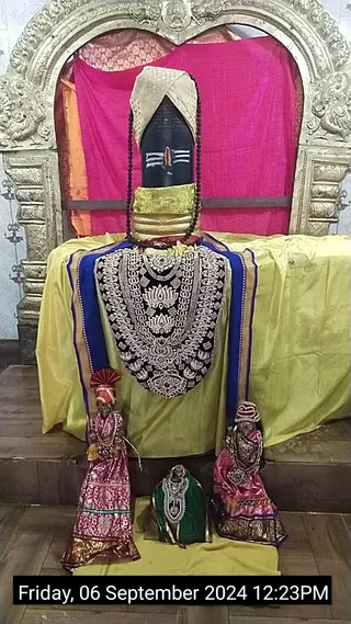 A photograph of a Shiva lingam deity murti dressed for worship inside an ornate gilded arched shrine with a magenta backdrop. The black lingam bears three horizontal white ash lines and is wrapped with a cream and gold cloth turban, a band of yellow-green brocade, and a dark rudraksha-style bead garland. A heavily embroidered black and silver garment with lotus motifs covers the body, edged in blue and gold. Two small figures in pink and gold sarees with decorated headpieces stand at the base on either side, and a small green-clad object with silver ornaments sits in front. A yellow cloth covers the platform. A digital timestamp reads Friday, 06 September 2024 12:23PM. No physical people are visible.