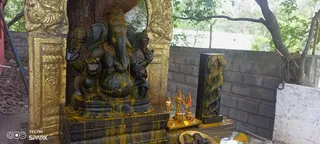 A photograph of a large dark stone Ganesha murti seated on a pedestal inside an ornate gold-painted arch, its surface smeared with yellow turmeric paste. Four arms are visible, with a modak-like sweet and a rope or goad held in the upper hands. Beside it stand smaller gold-coloured deity figures on a low platform with small offerings and a flower, and a dark carved snake-entwined pillar stands against a concrete block wall. A rudraksha or prayer-bead mala hangs from a branch at right, and trees and a hose are visible behind. No people are present.