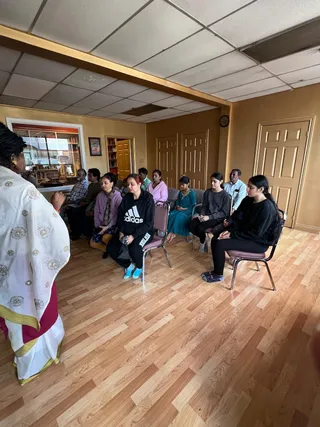 A photograph of a room with wood-effect laminate flooring, gold-painted walls and a drop ceiling, where a group of about ten people sits on folding chairs facing left. A woman standing at the left edge wears a white shawl embroidered with gold motifs over a white and maroon garment, and her hands are folded in front of her. The seated people include a woman in a black Adidas hoodie with blue shoes, women in pink and lavender tops, a woman in a teal salwar kameez, and young women in black sweatshirts. Several men sit at the back, one in a plaid shirt and another in a light shirt. Several sit with eyes closed as if in meditation. No a person candidate is visible.