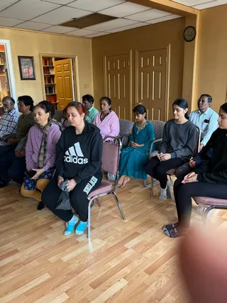 Indoor photograph of a group of about nine seated people in folding chairs on a wooden laminate floor in a room with mustard-yellow walls, a drop ceiling and a wall clock. Most have their eyes closed and hands resting in their laps. A woman in a black Adidas hoodie and black track pants sits in front holding a dark plastic bag and wears blue socks. A girl in a teal embroidered dress sits with hands clasped, and a teenager in a grey sweatshirt sits beside her. A woman in a pink top and a man in a light blue shirt sit further back, with a man in a plaid shirt at left. A bookshelf and an open door are visible at left, and a framed picture hangs on the wall.