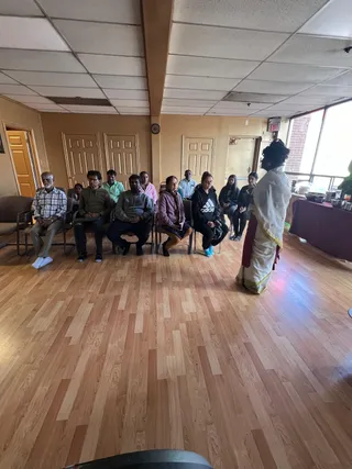 A photograph of a room with a wood-laminate floor and drop ceiling. A standing figure in a white and gold-bordered dhoti or veshti with a red sash faces a row of about a dozen seated people on chairs, including an older man in a checked shirt, several men in casual sweatshirts and shirts, women in a pink top, a dark Adidas hoodie and a purple sweater. A ritual table with a maroon cloth and metal vessels stands at the right. The standing figure's appearance is ambiguous and no clear a person cues are visible, so no identification is made.