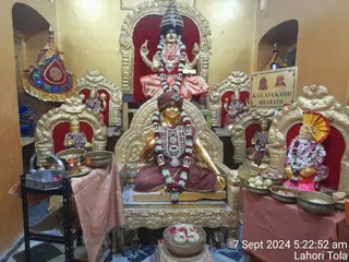 A shrine room with golden thrones and red velvet backs holds several seated murtis. At the centre, a seated golden figure wears a maroon garment, a maroon head wrap, a large ornate necklace and a long garland of white and green flowers, with one hand raised in blessing. Behind, a four-armed deity murti in pink and red garments, a crown and a long garland sits on a higher throne. To the right, a Ganesha murti with a yellow fan-shaped headdress and pink garland sits on a gold throne. Brass bowls with fruit and offerings stand on a draped table, and a stand with oil lamps and small dishes sits at left. A decorated umbrella-like canopy hangs at left. The timestamp reads 7 Sept 2024 at 5:22:52 am.