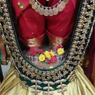 Close view of a deity murti from the waist down, dressed in a red silk sari with gold brocade borders and a gold-toned lower garment. A layered necklace of large round coin-like pendants hangs at the chest. Beneath the figure is a silver feet-shaped base with yellow and red roses and a yellow flower placed as offerings. The murti is framed by a green velvet panel decorated with silver-toned floral and leaf embellishments and round studs. No people are visible.