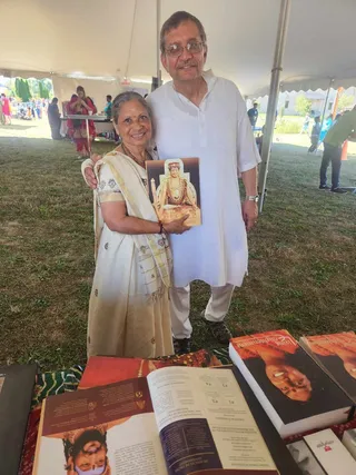 Outdoor booth under a white tent on grass at a festival. A smiling older woman in a cream and gold-bordered sari stands beside an older man in a white kurta and white trousers with glasses and a wristwatch. Both hold a printed booklet showing an ornate seated figure in gold jewellery and a red-orange backdrop. On the table in front are open books, one showing a red-toned portrait, another with a woman's face on the cover, and a book with a red-orange cover. Other attendees and tables are visible in the background under the tent.