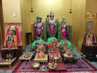 A shrine room with yellow walls and a gold-framed sign reading KAILASA SINGAPORE on the right. Three large deity murtis sit on a raised dais: the central figure wears a tall crown, a green-and-gold sari, a garland and a trident, with a snake over its head, while the flanking figures wear pink and green garments, a green turban and a pointed crown. Smaller murtis in orange and red stand below, with brass oil lamps, a kalasha pot, fruit offerings, a brass bell and a brass plate with lamps on a red carpet. A Happy Diwali banner hangs on the left wall. No people are visible.