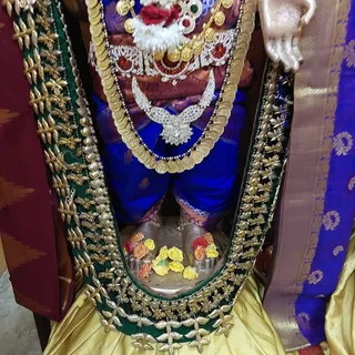 Close view of a decorated deity murti standing in a shrine, dressed in a blue garment with a maroon and gold brocade upper cloth and a large gold coin necklace with a jewelled silver-toned pendant. A red and white flower garland and red stones adorn the upper body. Its lower body is covered by a green velvet panel edged in gold and silver embroidery, above a silver base with yellow and orange roses placed at its feet. A purple and gold sari border hangs at the right. No people are visible.