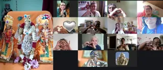 A video call grid of participants making heart hand gestures, shown beside a photograph of three decorated deity murtis on an orange cloth. The left murtis wear crowns and garlands with pink and purple flowers; the central figure is blue-grey with a peacock feather behind, and the right figure wears a colourful turban and necklaces. The grid has about a dozen small video tiles: women and a few men in home settings, some with bindis and rudraksha-style beads, one tile showing a stone Shiva statue, and a silver heart graphic. Several tiles show people in saffron or white clothing. The screen shows an online devotional gathering.