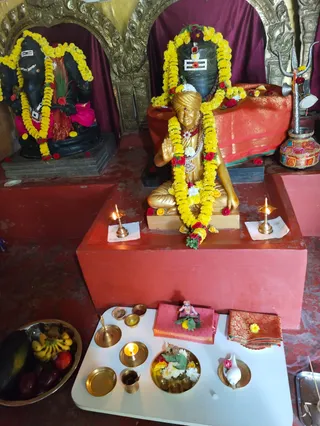 Photograph of a temple shrine with no living people. Centre: a gold-coloured seated murti of a figure with a saffron-coloured turban, hands in anjali, wearing a red and white beaded necklace and a red forehead mark, garlanded with yellow marigolds and red roses. Behind it a black stone Shivalinga with white tripundra lines, red and white flowers, and a silver trident stands on a red cloth. At left a black Ganesha murti in yellow marigold garlands. Two brass oil lamps with lit wicks sit on white paper on a red platform. In front, a white table holds brass plates with a lit ghee lamp, flowers, a conch shell, a brass kalasha, a small brass cup, and a bowl of fruit including bananas and apples. Red and gold silk cloths lie on the table.