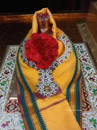 A photograph of a Shiva linga draped in a yellow cloth with a green and maroon zari border, adorned with a large jeweled necklace of white stones with green and pink accents and a central round stone, and topped with a large red carnation. The linga sits on a white and gold ornate patterned altar cloth with red and green floral motifs. No people are visible.
