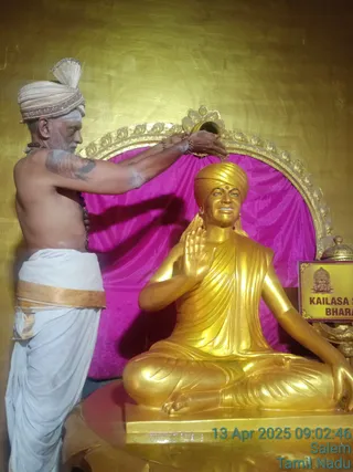 A photograph of a shirtless priest with a white-and-cream turban and a rudraksha mala, wearing a white dhoti with a gold-edged sash, pouring liquid from a small vessel over the head of a seated golden statue. The golden murti sits in a meditative pose with one hand raised in abhaya gesture, wearing a golden turban and robe, before a magenta cloth backdrop and an ornate golden peetha frame. A small plaque beside the statue reads KAILASA S... and BHARA..., and a timestamp in the corner reads 13 Apr 2025 09:02:46 Salem Tamil Nadu. The scene appears to show abhishekam of a statue. The statue is not a physical person.