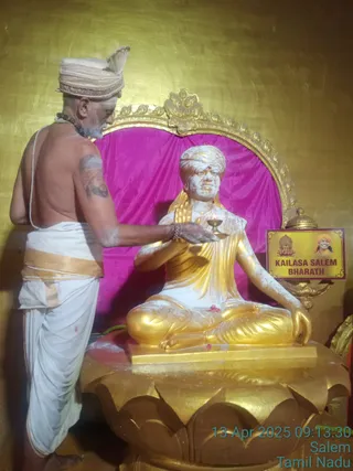 A bare-chested priest in white dhoti with a gold-bordered cloth, wearing a cream turban and a rudraksha mala, stands at left holding a small oil lamp over a seated golden statue. The golden statue sits in lotus pose on a lotus-petal pedestal before a magenta velvet backdrop framed in gold. The statue wears a golden robe and a cream turban, and its surface is dusted with white powder. A sign at right reads KAILASA SALEM BHARATH with a small portrait. A timestamp reads 13 Apr 2025 09:13:30 Salem Tamil Nadu. The scene shows an aarti offering performed before a deity-like statue.