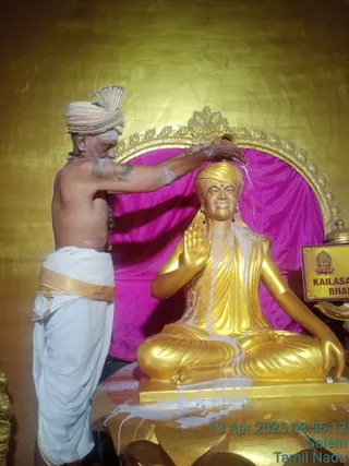A photograph of a priest performing abhishekam on a seated golden statue of a teacher in a meditation pose with raised right hand. The priest, shirtless with a white turban and white dhoti with a gold border, pours liquid over the statue's head with his right hand and holds a bead mala at his wrist. Milky liquid streams down the golden robes and base. Behind the statue is a magenta velvet backdrop framed by an ornate gold arch, and a sign reads KAILASA with partial text BHAR. The timestamp reads 13 Apr 2025 08:56:12 Salem Tamil Nadu. The statue itself is a depiction, not a living person, and the priest is a physically present person whose identity is not established by appearance.
