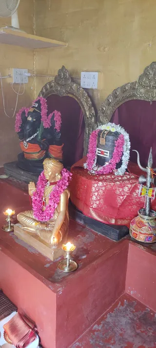 A shrine corner with three sacred images. On the left a black Ganesha murti with a pink flower garland and orange cloth. In the centre a golden seated statue of a man with a turban, pink flower garland, red bead necklace, hands raised in a greeting gesture, placed on a red pedestal. On the right a black Shiva lingam draped in a red and gold brocade cloth, wearing a pink and white flower garland, with a silver trident beside it and a decorated kalasha pot. Two brass oil lamps burn in front, and gold-framed arches and maroon drapery form the backdrop.