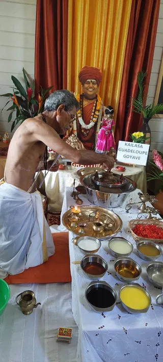 A photograph of an abhishekam ritual at a KAILASA centre. A bare-chested priest in white dhoti with orange-gold border sits on an orange cushion at left, pouring from a brass vessel over a large steel bowl on a white table. Behind him a framed portrait of a smiling figure wearing an orange turban, orange sari-like robe, marigold and white garlands and rudraksha mala stands on a draped stage with a white and gold cloth backdrop, flanked by potted tropical plants. A pink-clad doll-like figure and a brass lamp stand on the table, with steel bowls of milk, honey, ghee and other liquids, a brass plate with lit oil lamps and a basket of red flower petals in front.
