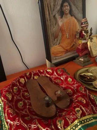 A photograph of a home shrine on an orange cloth. In the foreground, a pair of wooden padukas (sandals) with knob-handles rests on a red velvet cushion embroidered with gold sequin paisley motifs and green border trim. Behind them, a framed photograph shows a smiling figure in orange robes and a long white beaded mala, seated cross-legged before a banyan tree trunk with one hand raised in blessing; the framed image is a depiction. To the right, a small brass deity murti in a red and gold garment with a jeweled crown stands beside a brass tray holding sweets, and a framed yantra picture is partly visible at the far right. No people are physically present.