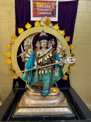 A large gilded metal murti of a multi-armed deity stands on a silver-toned pedestal set on a black platform in front of a purple velvet backdrop and a gold-painted wall. The deity wears a black turban with a red crest, teal and gold silk garments, layered gold necklaces, a rudraksha mala, and a cluster of brass bells at the waist. It holds a silver trident and a sword among its raised arms, and a small orange fruit sits near its hand and foot. A gold circular halo framed with yellow lotus petals surrounds the figure. Above, a framed sign reads KAILASA TORONTO CANADA with a small portrait of a smiling saffron-robed man. No physical people are visible.