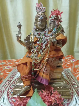 A brass deity murti seated on a brass pedestal, draped in orange and maroon silk cloth and layered with jeweled necklaces of red, green and white stones and gold chains. Pink ixora flowers adorn its crown and lie at its base, with a green leaf in the foreground. The murti sits on a red and gold embroidered cloth against a pale gold curtain. No people are visible.