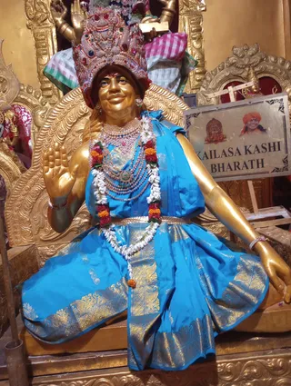 A gilded statue seated on an ornate golden throne, wearing an elaborate jeweled crown with red and green stones, layered gold and pearl necklaces, and a royal blue silk garment with gold brocade borders and a gold waist sash. A garland of white jasmine and orange marigolds hangs across its chest. The right hand is raised in blessing while the left rests on the lap. Behind it, a framed sign reads KAILASA KASHI BHARATH and shows a small deity image beside a portrait of a smiling figure in an orange turban. The statue is a murti-style depiction, not a living person, and no physical people are present.