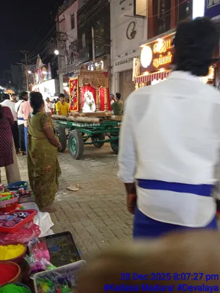 Night street scene in a busy Madurai market lane lit by shop lights and a lit sign. In the centre, a green flatbed cart with wooden poles carries a gilded wooden canopy with red drapes, under which a white-clad deity murti sits adorned with a green garland. A young man in a yellow shirt stands beside the cart, and another person in green stands on the cart near the canopy. On the left, a woman in a green sari stands by roadside stalls selling colourful plastic items and powders. In the foreground right, a man with a dark topknot wears a white shirt and blue dhoti-style cloth. Shop signs read G K Gem & Jewellery. The timestamp reads 28 Dec 2025 8:07:27 pm with #Kailasa Madurai #Devalaya.