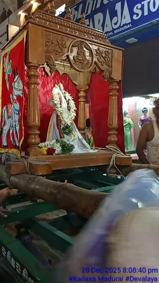 A wooden palanquin-style carriage with carved pillars and a carved arched top with a round medallion sits on a green wheeled wooden cart, draped in red cloth and a red-and-yellow banner painted with a white horse. Inside, a standing deity murti in white and lavender robes with a long white garland is decorated with white flower garlands and red flowers at its base. Blue sign text on a shop front behind and blurred foreground figures in a crowd are visible, with a green-and-pink dressed figure on a display at right. Timestamp in the lower right reads 28 Dec 2025 8:08:40 pm with the hashtag #Kailasa Madurai #Devalaya.